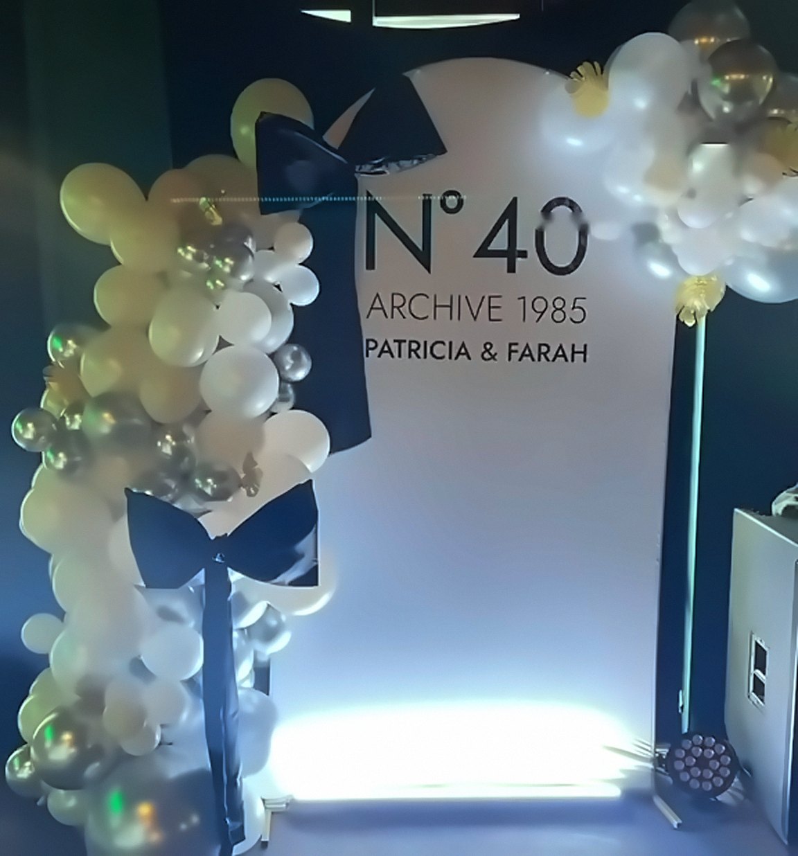 Fortieth birthday backdrop styled like a perfume bottle reading N°40 Archive 1985, with champagne, white and chrome balloon garlands and black satin bows